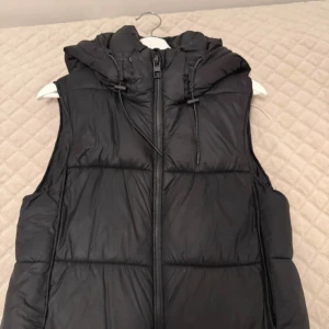 Svart dunväst från Zara med huva - Säljer en svart dunväst från Zara med huva och dragkedja framtill. Västen har en quiltad design och justerbar dragsko vid huvan. Perfekt att slänga över en hoodie för en chill look.