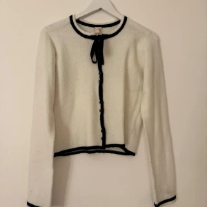 Vit kofta med svarta detaljer från Nelly - Supersöt vit kofta från Nelly i storlek XS med svarta kanter och knytband vid halsen. Koftan har knappar framtill och är långärmad med en croppad passform. Perfekt för dig som gillar klassisk stil med en twist.