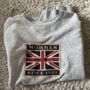 Grå sweatshirt från Morris - Säljer en grå sweatshirt från Morris med Union Jack-flagga och texten 'Morris Since 1968' på bröstet. Tröjan har rund halsringning och ribbade muddar. Perfekt för dig som gillar klassisk och stilren streetwear.