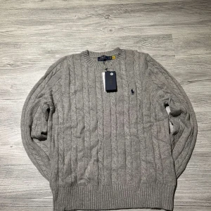 Grå kabelstickad tröja Polo Ralph Lauren - Kabelstickad grå tröja från Polo Ralph Lauren med klassisk rund halsringning och broderad logga på bröstet. Tröjan är i mjuk ullblandning och har ribbade muddar vid ärmslut och nederkant. Perfekt för lager-på-lager under kyliga dagar.