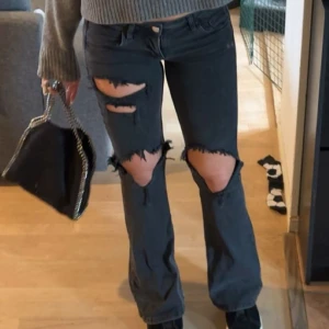 Svarta bootcut jeans med slitningar - Säljer ett par svarta jeans med bootcut-modell och stora slitningar över knäna och låren. Jeansen har klassisk femficksdesign och stängs med knapp och dragkedja. Perfekta för en edgy streetstyle-look.