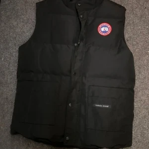 Svart dunväst från Canada Goose - Säljer en svart dunväst från Canada Goose med klassisk logga på bröstet och stora fickor framtill. Västen har knappar och dragkedja framtill och är perfekt för lager-på-lager under kyliga dagar. Stilren och enkel design som funkar till många outfits. Använd fåtal gånger!
