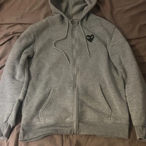 Grå Comme des Garçons Play hoodie - Grå hoodie från Comme des Garçons Play med dragkedja, huva och snörning. Ikoniskt svart hjärta med ögon på bröstet och stort hjärta på ryggen. Mjuk bomullskänsla och kängurufickor framtill. Perfekt för en chill och stilren look.