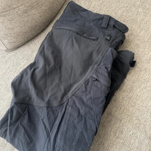 Svarta byxor från Haglöfs - Svarta cargopants från Haglöfs med flera praktiska fickor och förstärkta partier på knän och bak. Byxorna har dragkedja, bälteshällor och är tillverkade i slitstarkt material. Strl XL 
