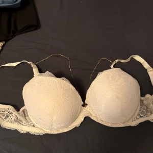 Vit spets-bh med guldkedja - Säljer en vit bh med vadderade kupor och vacker spetsdetalj över hela framsidan. Bh:n har en tunn guldfärgad kedja som dekoration mellan kuporna och justerbara axelband. Perfekt för dig som gillar unika detaljer och romantisk stil. Push up