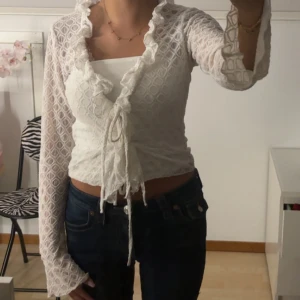 Vit omlottblus med volang och knyt - Säljer en vit omlottblus med volangkant längs halsringningen och knyt framtill. Blusen är långärmad och har ett genombrutet spetsmönster som ger en romantisk vibe. Croppad passform som visar midjan, perfekt att styla med höga jeans.