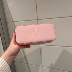 Pandora rosa smyckesväska med spegel - Supersöt och praktisk smyckesväska från Pandora i ljusrosa syntetmaterial. Väskan har guldfärgad dragkedja, spegel på insidan av locket (den är också uttagbar) och flera fack för ringar, örhängen och halsband. Även hållare för alla berlocker man har från Pandora. Sedan har den en liten kudde på plan 2 där man kan lägga sitt armband eller sitt favorit halsband!! Perfekt att ha med sig när du vill hålla koll på dina smycken. Köpt för ca 6 månader sedan, väldigt praktisk!! Kostar 449 kr på Pandora