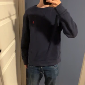 Mörkblå tröja från Polo Ralph Lauren - Snygg mörkblå tröja från Polo Ralph Lauren med klassisk röd logobrodering på bröstet. Tröjan har rund hals och långa ärmar, perfekt för dig som gillar stilren och enkel design. Passar dig som vill ha en chill och tidlös look.