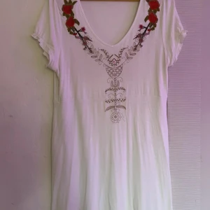 Vit blommig klänning från KappAhl XL - Vit kortklänning från KappAhl i storlek XL med v-ringning och broderade röda blommor vid halsen. Klänningen har ett unikt mönster framtill och korta ärmar. Perfekt för dig som gillar boho-vibbar och vill ha något bekvämt och snyggt.