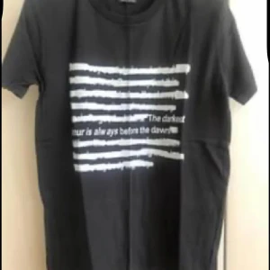 Svart t-shirt med vit text ny  - Svart t-shirt med korta ärmar och tryck framtill i form av vita streck och texten 'The darkest hour is always before the dawn'. Klassisk rund halsringning och rak passform. Perfekt för en chill och avslappnad stil.