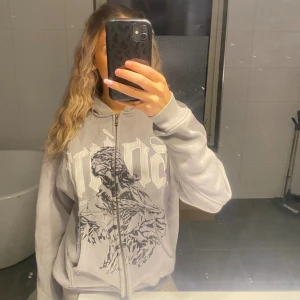 Grå hoodie med tryck och dragkedja - Säljer en grå hoodie med stor vit och svart tryckt design framtill. Tröjan har huva, hel dragkedja och ribbade muddar. Oversized passform och stora fickor framtill. Perfekt för dig som gillar streetstyle och vill ha något bekvämt och snyggt. Köpt för cirka 900 kr och använd max 5 gånger. Tror ej den går att köpa längre 