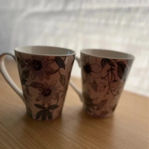 Två kaffemuggar - Bilderna visar två porslinsmuggar med blommigt mönster i ljusa färger. Muggarna har en elegant form och ser ut att vara i mycket gott skick utan synliga skador eller slitage. Perfekta för kaffe eller te och ger en stilren känsla till köket.