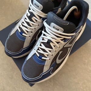 Dior B30 sneakers i blått och grått - Säljer ett par feta Dior sneakers med coola blå, grå och vita detaljer. Skorna har vit snörning, mesh och skinnpaneler samt en chunky sula. Märket syns tydligt både på plösen och sulan. Perfekta för dig som vill sticka ut med lyxig streetstyle. 7/10 Skick. Vid frågor kontakta mig. 
