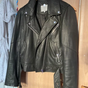 Svart skinnjacka från Dante 6 - Säljer en svart skinnjacka från Dante 6 med coola detaljer som dragkedjor, knappar och bälte i midjan. Jackan har en klassisk bikerstil med croppad passform och långa ärmar med dragkedjor vid ärmslut. Perfekt för dig som vill ha en edgy look. Motsvarar storlek S/xs. Ny pris ca 450€