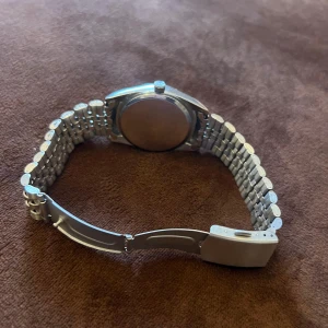 Silverfärgad quartzklocka med länkarmband - Stilren klocka med silverfärgat länkarmband och svart urtavla. Urtavlan har markerade index och datumvisning. Boetten är rund med räfflad kant och armbandet har viklås. Perfekt accessoar för dig som gillar klassisk design.