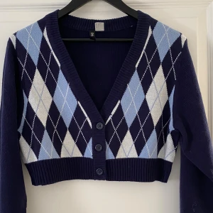 Mörkblå rutig kofta från Divided - Superfin croppad kofta från Divided i mörkblått med klassiskt argylemönster i ljusblått och vitt. Koftan har v-ringning, långa ärmar och knäppning med tre knappar framtill. Perfekt för lager-på-lager och snygg till jeans eller kjol.