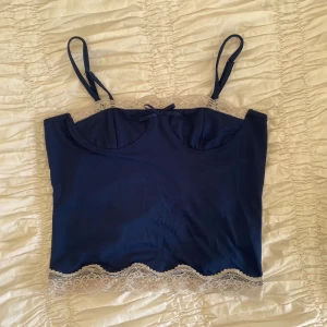 Mörkblå linnetopp med spetsdetaljer - Supersöt mörkblå linnetopp med justerbara smala axelband och vit spets längs både halsringning och nederkant. Toppen har en liten rosett framtill och är croppad i modellen 