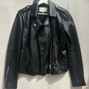 Svart skinnjacka från Vila - Snygg svart skinnjacka från Vila med klassisk bikerstil. Jackan har silvriga dragkedjor framtill och vid ärmsluten, kortare i modellen och i toppskick! 