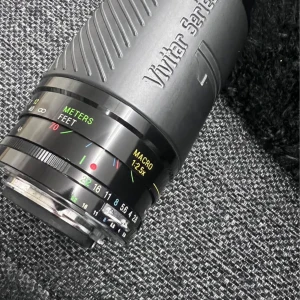 Vivitar Series 1 70-210mm f/2.8-4.0 Macro Zoom Lens - Vivitar Series 1 70-210mm f/2.8-4.0 macro zoomobjektiv med manuell fokus. Linsen har tydliga markeringar, macro 1:2.5x, och levereras med skyddspåse. Glaset ser klart ut och objektivet är i gott skick utan synliga repor eller skador.