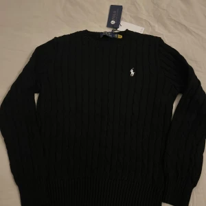 Svart kabelstickad tröja Polo Ralph Lauren - Svart kabelstickad tröja från Polo Ralph Lauren med klassisk rund hals och vit broderad logga på bröstet. Tröjan har långa ärmar och ribbade muddar vid ärmslut och nederkant. Perfekt för dig som gillar stilrena och tidlösa plagg.