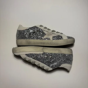  Golden Goose Glittriga - Säljer ett par GGDB sneakers med silverglittrig ovandel, grå mockadetaljer och stjärna på sidan. Skorna har grå snörning, vit sula och GGDB-logga på plösen. Perfekta för dig som vill sticka ut med en cool och trendig look.