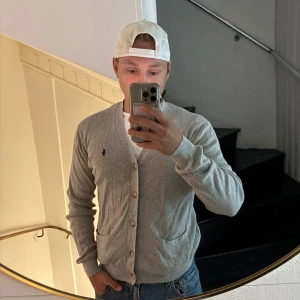 Grå kofta från Polo Ralph Lauren M - Snygg ljusgrå kofta från Polo Ralph Lauren med klassisk broderad logga på bröstet. Koftan har v-ringning, knäppning framtill med fyra knappar och två fickor. Tillverkad i mjuk bomull, perfekt för lager-på-lager-stil.