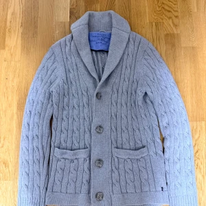 Grå kabelstickad kofta från GANT - Snygg grå kabelstickad kofta från GANT med stora knappar framtill och två fickor. Koftan har en bred krage och långärmade ribbade muddar. Perfekt för dig som gillar klassisk stil med en modern twist.