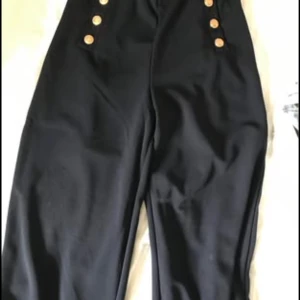 Blåa vida byxor med guldknappar - Snygga blå byxor med hög midja och vida ben. Byxorna har dekorativa guldknappar framtill på båda sidor som ger en cool och trendig look. Perfekta för dig som gillar statement-plagg och vill sticka ut lite extra.