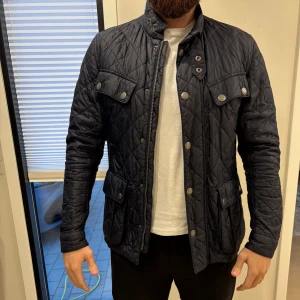 Marinblå quiltad jacka med fickor - Barbour  - Klassisk quiltad jacka från Barbour 🧥✨ Färg: marinblå Storlek: S  En tidlös modell med snyggt quiltat mönster, praktiska fickor och hög krage med tryckknappar. Perfekt för vår och höst – både stilren och bekväm.  Jackan har ett litet hål i armen som går att sy. I övrigt bra skick. Se bilder. 