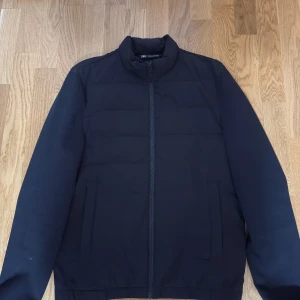 Svart pufferjacka från Zara - Stilren svart pufferjacka från Zara med hög krage och dragkedja framtill. Jackan har två diskreta fickor på sidorna och quiltad design som ger en clean look. Perfekt för kyligare dagar och enkel att matcha med olika outfits.