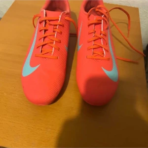 Nike fotbollsskor i neonrosa - Säljer ett par Nike fotbollsskor i neonrosa med ljusblå swoosh och detaljer. Skorna har snörning och är tillverkade i syntetmaterial med låg profil och platt sula för bra grepp på planen. Perfekta för dig som vill sticka ut på matchen!