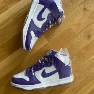Nike Dunk High Purple/White - Säljer ett par Nike Dunk High sneakers i vitt och lila. Skorna har klassisk hög siluett, snörning och den ikoniska swoosh-loggan på sidorna. Ovandelen är i skinn med perforerad tåbox för extra andningsförmåga. Perfekta för dig som vill sticka ut med färg på fötterna.
