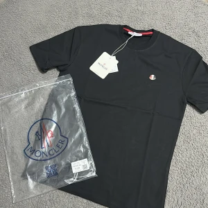 Svart Moncler t-shirt med logga - Diskret Moncler-logga i rött, vitt och blått på bröstet. T-shirten har en stilren look och är tillverkad i mjuk bomull. Perfekt för dig som gillar exklusiva märken och enkel design. Den kanske ser lite smutsig ut men det är bara kameran, den är helt ny🤟