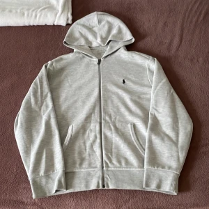 Ljusgrå hoodie från Polo Ralph Lauren - Ljusgrå hoodie från Polo Ralph Lauren med dragkedja framtill och klassisk svart logga på bröstet. Tröjan har huva, två fickor och är tillverkad i mjuk bomullsblandning. Perfekt för en chill och stilren look.