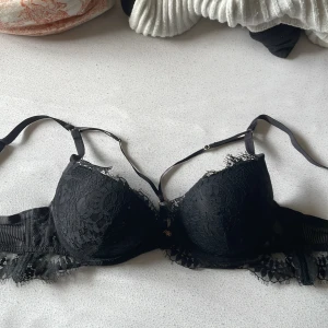 Svart spets bh med detaljer - Säljer en svart korsett från Chantelle med snygga spetsdetaljer och tunna justerbara axelband. Kuporna är dekorerade med blommig spets och har en liten guldfärgad berlock framtill. Passar perfekt under en skjorta eller som statement-piece.