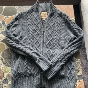 Grå stickad kofta från North Coast - Mysig grå stickad kofta från North Coast med grovt kabelstickat mönster och dragkedja framtill. Hög krage och ribbade muddar ger en snygg och avslappnad look. Perfekt för lager-på-lager under kyliga dagar.