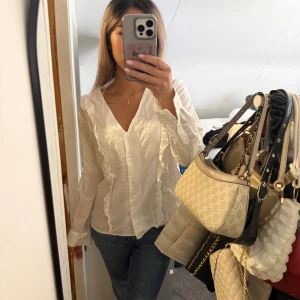 Vit volangskjorta med v-ringning - Säljer en vit skjorta med v-ringning och snygga volangdetaljer längs framsidan. Skjortan har långa ärmar och knappar framtill. Perfekt för dig som gillar en feminin och trendig look. Materialet känns lätt och luftigt. Står ingen storlek men passa mig som bruka ha s