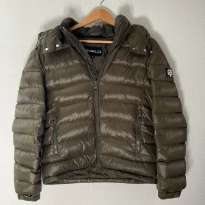 Olivgrön pufferjacka från Bvaldi - Säljer en olivgrön pufferjacka från Bvaldi med huva och två dragkedjeförsedda fickor. Jackan har quiltad design och en patch på ärmen. Perfekt för kyliga dagar och har en snygg, modern look.