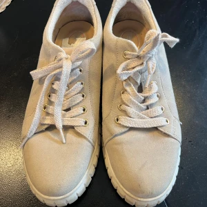 Beige sneakers från Tamaris i mocka - Snygga beige sneakers med chunky vit sula och ovandel i mjuk mocka. Skorna har rund tå, snörning med grova grå (och glittriga) skosnören och guldfärgade öljetter. Perfekta för dig som gillar en clean och stilren look med lite extra höjd i sulan.Använda några gånger och har ett litet märke på vänster sko men inget som märks