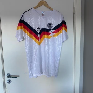 Tysklands retro fotbollströja Adidas L - Säljer en klassisk Tyskland fotbollströja från Adidas i storlek L. Tröjan är vit med ikoniska svarta, röda och gula V-ränder över bröstet, broderad landslagslogga och Adidas-tryck. Tillverkad i lätt och glansigt material, perfekt för fotboll eller samling.