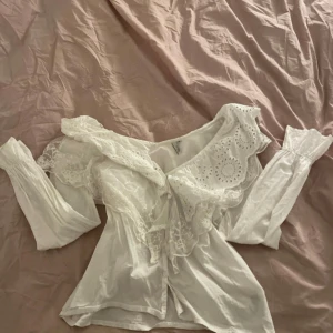 Vit blus med broderade spetsdetaljer - Superfin vit blus med långa ärmar och bred volangkrage med broderade hålmönster och spets. Blusen har omlottkänsla och knyt framtill, samt romantiska detaljer som ger en vintagevibe. Perfekt för dig som gillar en stockholmsstil 