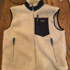 Beige fleeceväst från Patagonia - Säljer en beige fleeceväst från Patagonia med hög krage och dragkedja framtill. Västen har en mörkblå bröstficka med dragkedja och två sidofickor med blå detaljer. Perfekt för lager-på-lager och outdoor vibes.