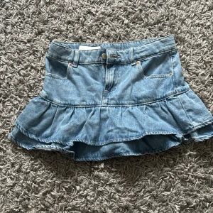 Blå jeanskjol med volang - Supersöt blå jeanskjol med volangkant och klassiska fickor fram. Kjolen har en kort modell och är tillverkad i denim med bälteshällor och knappstängning. Perfekt för dig som gillar en trendig och avslappnad stil.