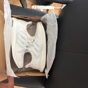 Vita Adidas Ultraboost sneakers - Säljer ett par fräscha Adidas Ultraboost sneakers i helvitt med stickad ovandel och klassiska tre ränder på sidorna. Skorna har snörning, platt sula och en bekväm boost-mellansula. Perfekta för dig som gillar stilrena och sportiga sneakers.