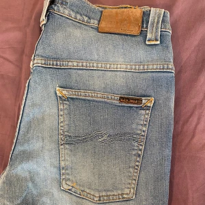 Ljusblå jeans från Nudie Jeans Co - Säljer ett par ljusblå jeans från Nudie Jeans Co i ekologisk bomull. Klassisk femficksmodell med snygga slitningar och kontrastsömmar. Jeansen har en rak passform och normal midja, perfekta för en avslappnad stil.