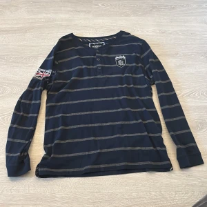 Randig långärmad tröja Bondelid XL - Mörkblå långärmad tröja från Bondelid i storlek XL. Tröjan har gråa ränder, knappar vid halsen och broderad logga på bröstet samt patch med brittisk flagga på ärmen. Skön bomullskänsla och klassisk stil.