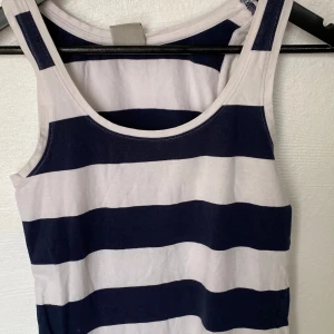 Randig linnetopp från Tribute - Säljer en snygg randig linnetopp från Tribute i storlek M. Toppen har breda marinblå och vita ränder, rund halsringning och ärmlös design. Perfekt för en avslappnad och cool stil. Tillverkad i mjuk bomull som känns skön mot huden.