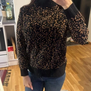 Svart leopardmönstrad stickad tröja - Säljer en svart stickad tröja med brunt leopardmönster och hög krage. Tröjan har långa ärmar och ribbade muddar vid ärmslut och nederkant. Perfekt för dig som vill ha en trendig och bekväm look.