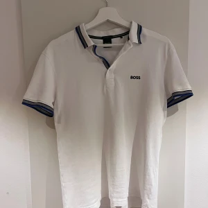 Vit pikétröja från BOSS, storlek S - Vit pikétröja från BOSS med blå och svarta detaljer på krage och ärmslut. Klassisk polokrage och kort ärm, broderad BOSS-logga på bröstet. Tillverkad i bomull för en skön och luftig känsla. Perfekt till jeans eller shorts. Inga fläckar eller några tecken på användning!