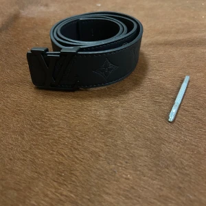 Svart Louis Vuitton skärp i skinn - Snyggt svart skärp från Louis Vuitton i skinn med präglad monogramdesign och mattsvart LV-spänne. Bältet har en stilren look med diskreta detaljer och passar perfekt för dig som vill ha något exklusivt och trendigt.